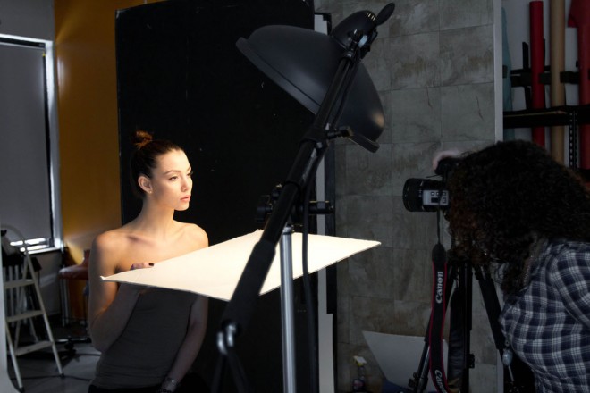 A picture of a model holding a reflector in her lap. A light over the camera shines down and the light is reflected back up into her face.