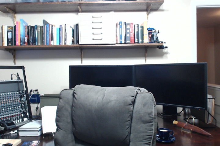 A view of a desktop containing two monitors and a sound mixer. Shelves are in the background.